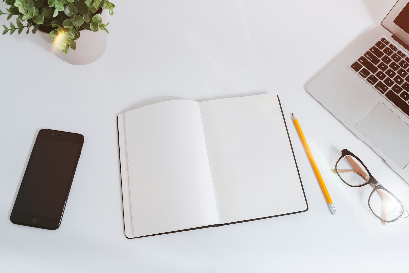 Caderno aberto, notebook e celular sobre uma mesa de trabalho.